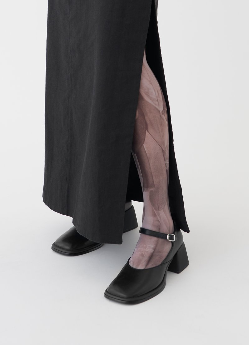 Chaussures à talons en cuir noir avec un bout carré, dotées d'une sangle et d'un talon carré, associées à des collants transparents à motifs et à une longue jupe noire.