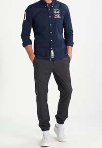 Man med marinblå skjorta med broderade logotyper, mörkgrå joggerbyxor, vita sneakers, händerna i fickorna, stående framför vit bakgrund.