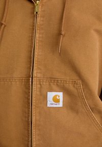 Haut à zip marron avec cordons et un patch logo Carhartt blanc sur la poche avant, montrant la texture du tissu et les détails de couture.