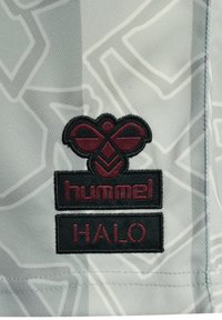 Hummel Halo-märket har en svart broderad logotyp med en maroon-färgad bikupeliknande design på en ljusgrå, texturerad tyg med subtila geometriska mönster.