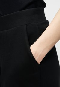 Χέρι χωμένο στην τσέπη μαύρου ψηλόμεσου παντελονιού που φοράει με μαύρο τοπ, δείχνοντας λεία ύφανση και απλό σχέδιο.