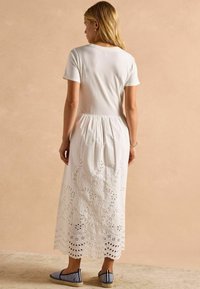 Weißes Baumwollkleid mit einem taillierten Oberteil und einem fließenden Rock, der am Saum Spitzenstickerei aufweist. Kombiniert mit blau gestreiften Espadrilles.