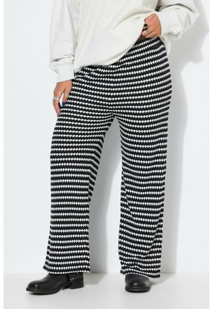 Persona con pantalones de patrón en zigzag blanco y negro, botas negras, una camiseta de manga larga de color claro y anillos en los dedos.