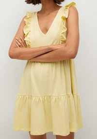 Žluté šaty vyrobené z jemné látky s rozšířenou sukní, s volánkovými okraji na rukávech a véčkovým výstřihem.
