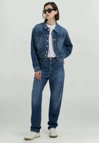 Jaqueta de ganga e jeans de cintura alta em azul médio. A jaqueta possui dois bolsos frontais, uma t-shirt branca por baixo e ténis brancos.