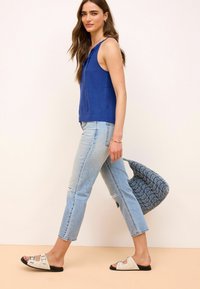 Blå ærmeløs top, lyseblå slidte jeans, beige sandaler med sort sål og en blå mønstret håndtaske, går på en neutral baggrund.