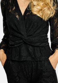 Melna mežģīņu blūze ar garām piedurknēm, V veida izgriezumu un savītu priekšpusi. Visa blūze ar teksturētu ziedu rakstu, pepļuma siluets un kabatas.