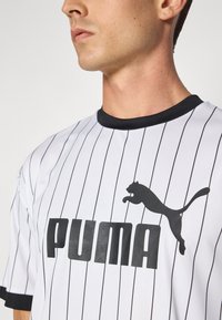 Hvidstribet trøje med sort krave og ærmeaccenter, der har et sort Puma-logo og fed "PUMA" tekst. Fremstillet i glat stof.