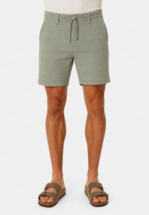 Mann trägt hellgrüne strukturierte Shorts mit Kordelzug, weißes geripptes Shirt und braune Sandalen mit Schnalle, steht vor einem schlichten Hintergrund.