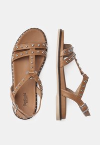 Sandali in pelle marrone con accenti di borchie, cinture incrociate, suola piatta e chiusura a fibbia per una vestibilità regolabile. Suola in gomma antiscivolo.