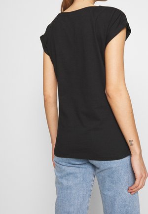 Kobieta ubrana w prostą czarną koszulkę z krótkim rękawem i jasnoniebieskie jeansy, pokazująca tylny widok z małym tatuażem litery "E" na lewym przedramieniu.