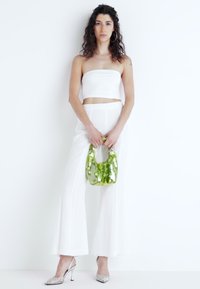 Biely crop top bez ramienok, široké nohavice, strieborné vysoké topánky na podpätku a zelená lesklá kabelka s jedinečným tvarom a textúrou.