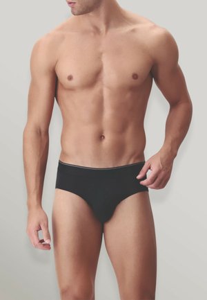 Uomo muscoloso in mutande nere in piedi contro uno sfondo chiaro e uniforme, con il torso e le cosce superiori visibili.