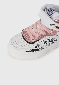 Baskets blanches avec des accents métalliques roses, des motifs graphiques noirs, un bout perforé et une sangle en velcro sur un laçage rose.
