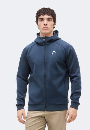 Mladý muž v tmavomodrej mikine na zips s kapucňou a béžových nohaviciach, stojaci na jednoliatom svetlom pozadí, čelom dopredu.