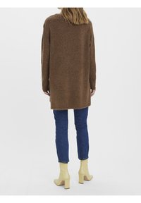 Brązowy sweter z dzianiny o luźnym kroju, z długimi rękawami i rozcięciami po bokach, zestawiony z niebieskimi dżinsami i żółtymi botkami na obcasie.