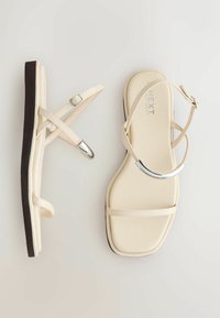 Bézs színű lapos szandál vékony pántokkal, ezüst díszítéssel az elülső pánton, állítható boka csattal és látható fekete talpélekkel.