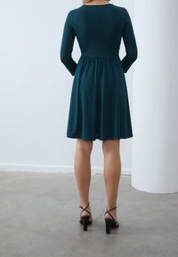 Långärmad teal klänning med en figursydd överdel och utsvängd kjol, med rund halsringning och mjukt, lätt tyg. Klacksandaler.