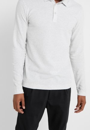 Homme portant un polo à manches longues gris clair et un pantalon noir, debout les bras détendus le long du corps sur un fond blanc uni.