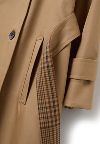Trinchera beige con una textura suave, que presenta un acento de cuadro de plaid marrón en el interior y un cierre de botón negro único.