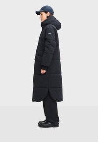 Čierny vystúžený kabát s kapucňou, s vlnitým lemom, bočnými vreckami a detailom loga na ramene, skombinovaný s tmavými nohavicami a čiernou obuvou.