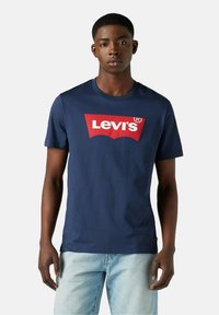 Hombre vistiendo una camiseta de Levi's azul marino con un logo rojo y blanco, combinada con unos pantalones vaqueros de mezclilla azul claro, de pie frente a un fondo liso.