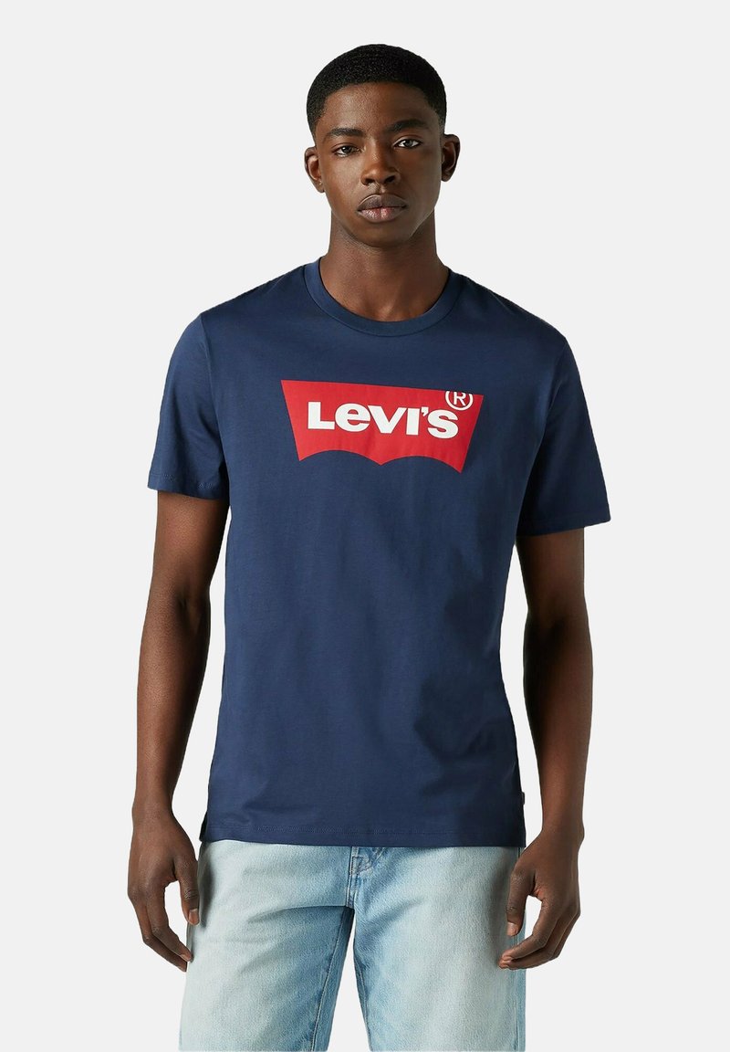 Hombre vistiendo una camiseta de Levi's azul marino con un logo rojo y blanco, combinada con unos pantalones vaqueros de mezclilla azul claro, de pie frente a un fondo liso.