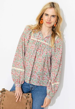 Femme blonde portant un chemisier à manches longues à motifs floraux, un jean bleu, des boucles d’oreilles orange, tenant un sac à main en cuir beige sur fond blanc.