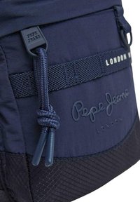 Zaino blu navy con tessuto testurizzato, dettagli intrecciati e una toppa in gomma con la scritta "Pepe Jeans London." Dotato di chiusura a coulisse.