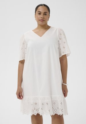 Femeie purtând o rochie albă cu decolteu în V, cu mâneci și tiv din dantelă cu flori decupate, stând în fața unui fundal simplu și deschis la culoare.