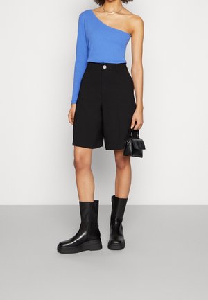 Žena v modrém topu s jedním ramenem a dlouhým rukávem, černé kraťasy po kolena, černé boty ke střední části lýtek, drží malou černou kabelku.