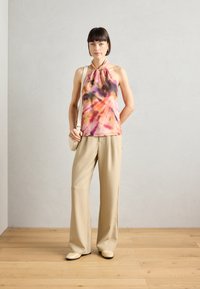 Ärmelloses pink-orange Batik-Top mit geknotetem Hals, kombiniert mit beigen weiten Hosen und cremfarbenen Schuhen. Holzfußboden im Hintergrund.