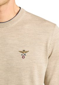 Maglione beige lavorato a maglia con scollo rotondo a costine, caratterizzato da un logo con ali dorate ricamate e dettagli in rosso e blu.