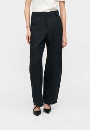 Person trägt hochtaillierte schwarze weite Hosen, ein weißes, eingestecktes Oberteil, schwarze Slingback-Schuhe mit spitzem Zehenbereich und ein Armband am linken Handgelenk.