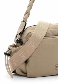 Sac en toile beige à bandoulière avec sangle tressée, poche zippée et détail de logo sur la sangle. Texture douce et design rectangulaire compact.