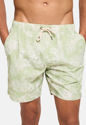 Bañador corto verde claro con estampado blanco de piñas tropicales y hojas de palma, cintura con cordón, llevado por una persona de pie con las manos en los bolsillos.