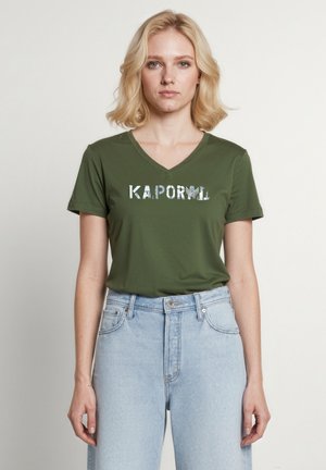 Mujer rubia de pie mirando hacia adelante, con una camiseta verde oliva con el logo de Kaporal y vaqueros de tiro alto en azul claro sobre un fondo liso.