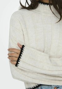 Femme en pull côtelé crème avec bordure noire aux poignets, les bras croisés sur un jean bleu clair.