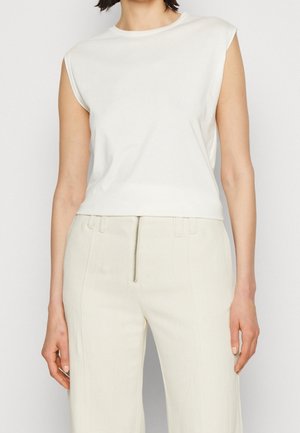 Frau trägt ein ärmelloses weißes Oberteil mit gepolsterten Schultern, kombiniert mit hoch taillierten weißen Hosen mit einem Reißverschluss vorne.