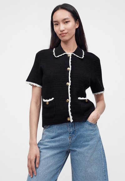 Jeune femme portant un haut en maille noir à manches courtes avec bordure blanche et boutons dorés, associé à un jean taille haute bleu clair.
