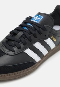 Zapatilla de cuero negro con rayas blancas, detalles de ante negro texturizado, cordones negros, suela de goma y logotipo azul de Adidas en la lengüeta.