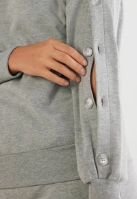 Grå sweatshirt med rund halsudskæring, ribbestrikkede ærmer og en knappet sideslids med fire store, lysegrå knapper. Blød tekstur.