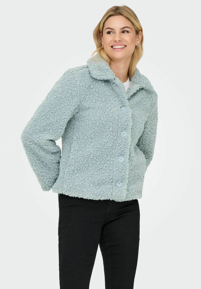 Light blue shearling jacket with a collar, button front, and side pockets; soft texture and cropped design. Worn with black pants.