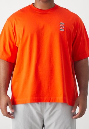 Mand iført en skrigorange kortærmet t-shirt med tre små broderede krokodillelogoer på venstre bryst og lysegrå bukser.