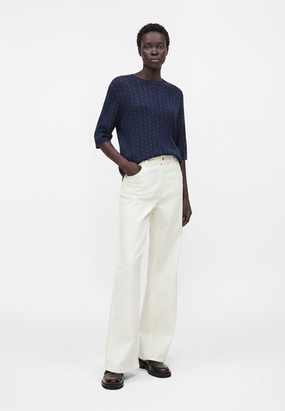 Γυναίκα όρθια με το ένα χέρι στην τσέπη, φοράει μπλε πουά πουλόβερ, λευκό παντελόνι με φαρδιά πόδια και σκούρα καφέ παπούτσια.