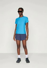 Moteris stovi, vilkėdama ryškiai mėlyną marškinėlį, tamsias šortus, baltas kojines, juodus sportinius batelius ir didelius juodus akinius nuo saulės, prieš paprastą foną.