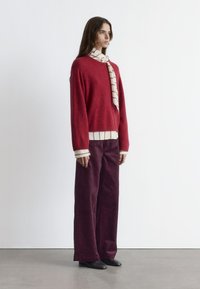 Red sweater with long sleeves, layered over a white striped shirt. Dark burgundy wide-leg corduroy pants and black footwear complete the look.