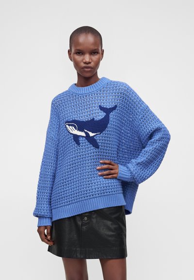 Femme portant un pull tricoté bleu avec un grand motif de baleine bleu foncé et blanc et une jupe en cuir noire, posant la main sur la hanche.