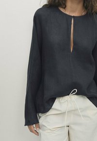 Blusa di lino blu navy con scollatura rotonda, dettaglio con apertura a chiave sul davanti e maniche lunghe, abbinata a pantaloni beige chiaro con cordino.