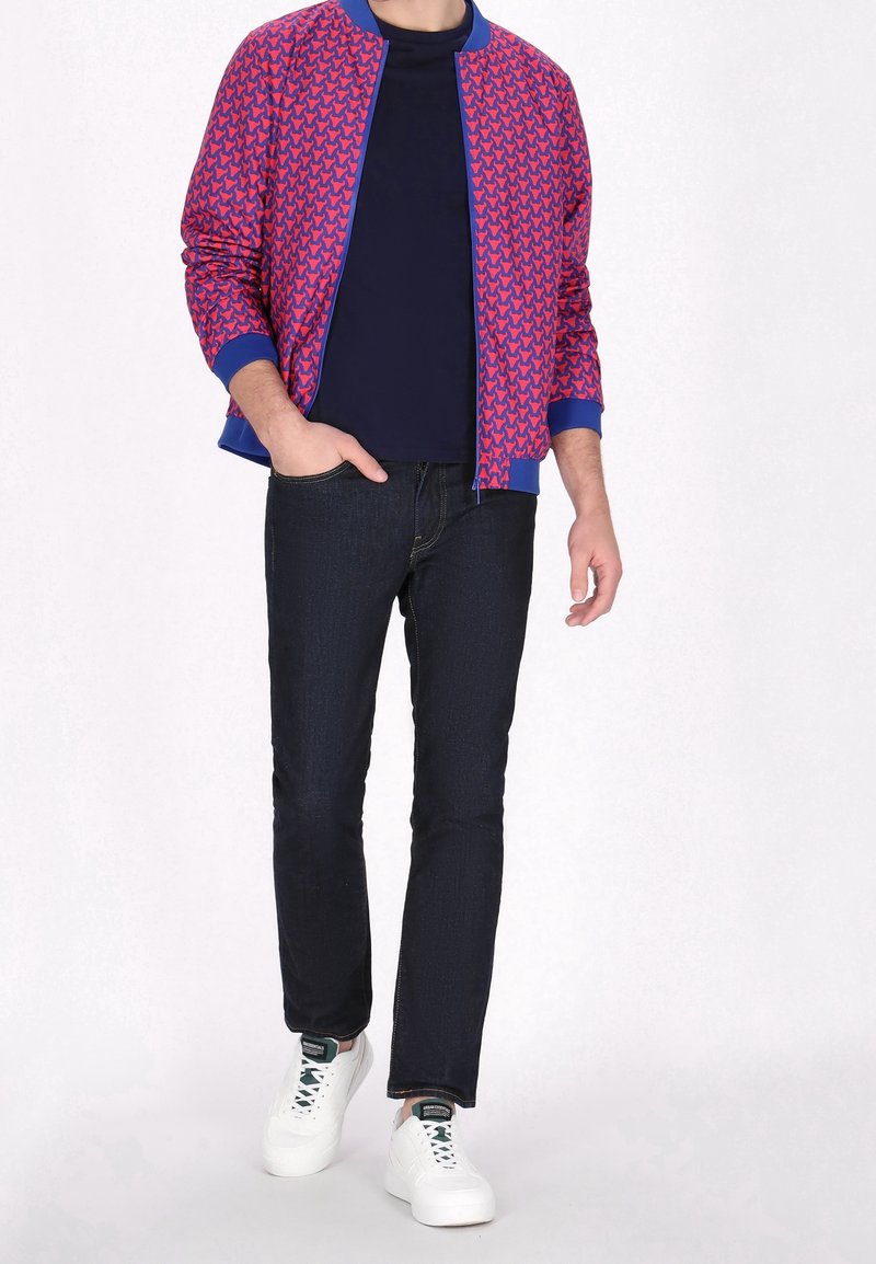 Homme portant une veste bomber rouge et bleue à motifs, chemise noire, jean foncé et baskets blanches, debout avec une main dans la poche.